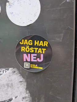 Street sticker The sticker is circular with a dark, slightly reflective background. The main text "JAG HAR RÖSTAT" is prominently displayed in large, bold yellow capital letters. Below this, the word "NEJ" is even larger, in bold purple capital letters with a subtle red drop shadow. In the bottom left quadrant, there is a small QR code with a purple outline. To the right of the QR code, in smaller yellow capital letters, is the phrase "OLYDIG FOLKOMRÖSTNING". The sticker appears to be adhered to a textured, dark grey surface, showing minor wear and tear.