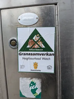 Street sticker A rectangular white sticker with a thin green border, applied to a metallic surface. The upper half features a large green triangle containing two cartoon animals: a brown rat-like creature on the left and a lighter brown monkey-like creature on the right, both appearing in a defensive or aggressive stance. Above the monkey, inside the triangle, is the text 'DEFEND SÖDERORT'. Below the triangle, in large bold black letters, is 'Grannsamverkan', followed by 'Neighbourhood Watch' in a smaller font. The bottom left corner has 'Stockholm', 'Södermalm', and 'Söderort' stacked. In the bottom center, there's a yellow crest with a crowned head, and 'Hammarby' written below it. The bottom right corner features a small logo with 'DEFEND SÖDERORT' and a silhouette of an assault rifle. The sticker shows some wrinkles and slight wear, indicating it's a physical sticker.