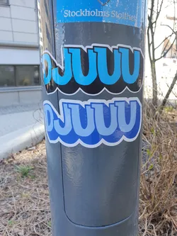 A blue and black sticker with repeating text. The text appears to be stylized and possibly nonsensical. The sticker is attached to a gray pole outdoors.