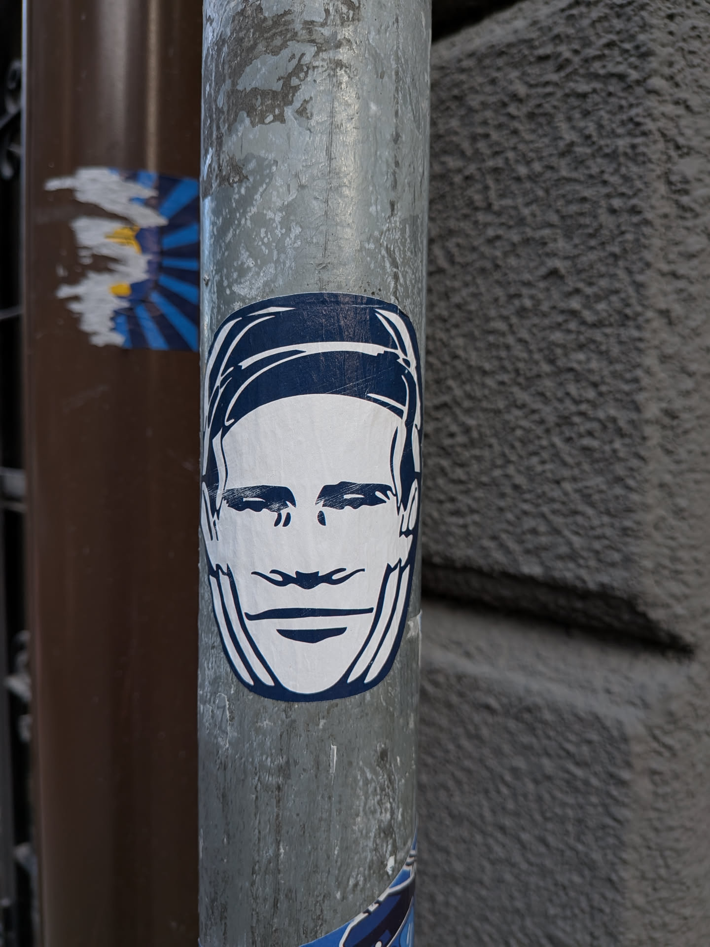 The sticker is centrally located on a weathered metal pole. It features a minimalist illustration of a face rendered in stark white against a deep blue outline. The face has simple, blocky features: two horizontal lines for eyes, a curved line for a nose, and a wide, open smile. The outline of the face and hair is in a dark blue, with some white space within the outline for the features. The sticker appears to be made of a matte material.