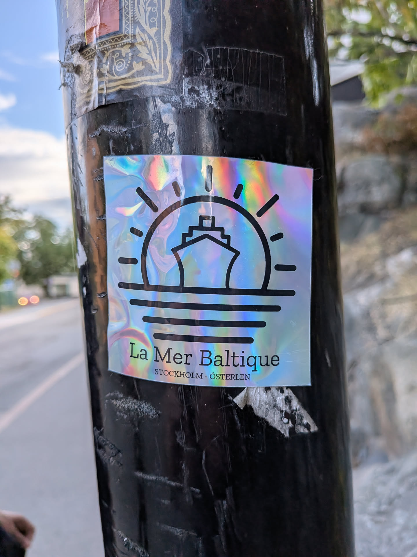 A square holographic sticker with a rainbow sheen. It displays a black minimalist icon of a ship sailing in front of a rising sun over water. Below, the text reads 'La Mer Baltique' and 'STOCKHOLM - ÖSTERLEN'.