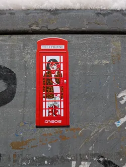 Street sticker The sticker is shaped like a traditional red British telephone booth. Inside, a stylized figure of a girl with dark hair is depicted, featuring a prominent red lightning bolt across her face, reminiscent of David Bowie's Aladdin Sane persona. She wears a brown and black leopard print coat over a light-colored garment and red cowboy boots adorned with white stars. She holds a red, heart-shaped telephone receiver to her ear. The top of the booth reads 'TELEPHONE', and 'PULL' is visible near the door handle. The overall aesthetic is bold and graphic, with a vibrant red dominating the design.