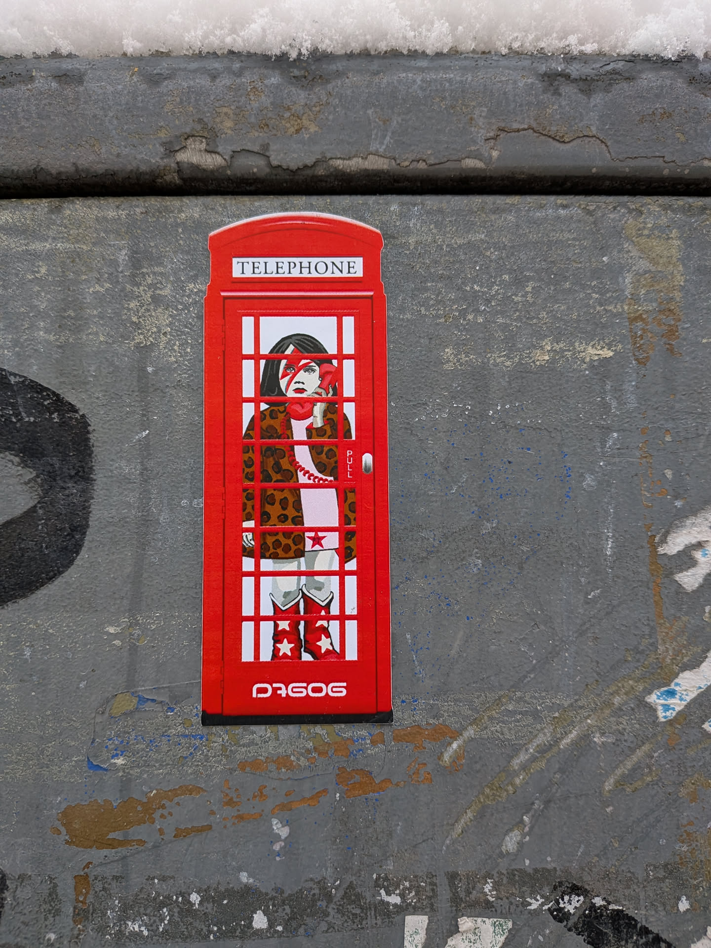 Street sticker The sticker is shaped like a traditional red British telephone booth. Inside, a stylized figure of a girl with dark hair is depicted, featuring a prominent red lightning bolt across her face, reminiscent of David Bowie's Aladdin Sane persona. She wears a brown and black leopard print coat over a light-colored garment and red cowboy boots adorned with white stars. She holds a red, heart-shaped telephone receiver to her ear. The top of the booth reads 'TELEPHONE', and 'PULL' is visible near the door handle. The overall aesthetic is bold and graphic, with a vibrant red dominating the design.