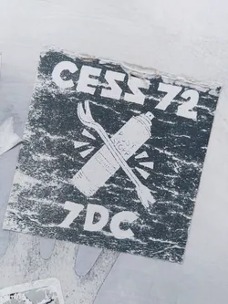 A dark grey sticker featuring the text "CESS72" at the top and "7DC" at the bottom.  A graphic of a spray can and a crowbar are crossed in the center. The style is reminiscent of graffiti art.