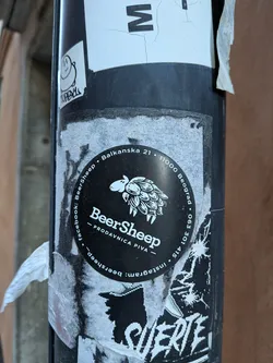 Street sticker A circular black sticker featuring a white stylized illustration of a sheep whose fleece is composed of hop cones, facing left. Below the illustration, the word 'BeerSheep' is prominently displayed in white, with '— PRODAVNICA PIVA —' (Beer Shop) underneath. Around the perimeter, in white text, are contact details and social media handles: 'Balkanska 21 • 11000 Beograd • 063 301 415 • instagram: beersheep • facebook: BeerSheep'. The sticker has a clean, monochrome design, standing out against the worn pole.