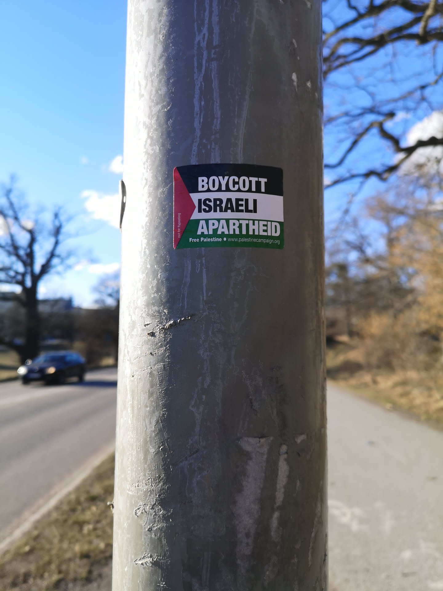 A rectangular sticker affixed to a grey metal pole. The sticker features a bold, multi-colored design. On the left side, a vertical strip displays the colors of the Palestinian flag: a red triangle on the left, followed by horizontal stripes of black, white, and green. The main body of the sticker contains text in a block format. The top line reads 'BOYCOTT' in white letters on a black background. The middle line reads 'ISRAELI' in black letters on a white background. The bottom line reads 'APARTHEID' in white letters on a green background. Below this, in smaller white text on the green background, it states 'Free Palestine' followed by a small white dot and 'www.palestinecampaign.org'. There is also very small, rotated text on the red triangle portion, which appears to be a disclaimer.