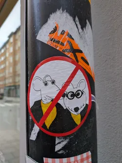 Street sticker A circular sticker featuring a red prohibition symbol (a circle with a diagonal line) over two cartoon mice. Both mice are white with black outlines, wearing black jackets or suits and yellow scarves or ties. The mouse on the right wears round black glasses and has prominent front teeth. The background within the red circle is white. The sticker has a hand-drawn, somewhat whimsical style.