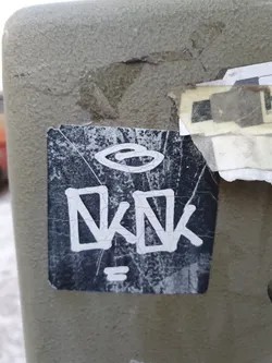 A worn black and white sticker is affixed to a light gray surface. The sticker features a stylized logo at the top and the letters "KNK" in a bold, slightly distorted sans-serif font at the center.  The letters appear to be graffiti-style. A small symbol resembling an equals sign is beneath the letters. The sticker shows signs of wear and tear, with some peeling and damage to the edges.
