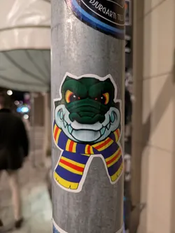 Street sticker A sticker featuring a grinning alligator wearing a scarf with blue, yellow, and red stripes. The gator has bright yellow eyes and a menacing look.