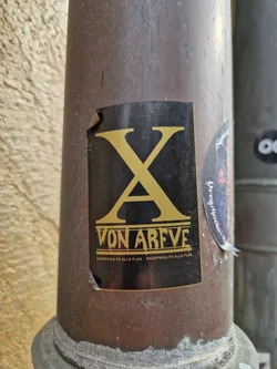 Street sticker A black sticker with gold lettering on a dark brown pipe. The sticker features a large stylized 'XA' logo, which is likely an abbreviation or a brand logo. Underneath the logo, the text 'VON ARFVE' is displayed, which could be a name, a brand, or a title. Repeated twice below that, there is the text 'SKARPNING PÅ ALLA PLAN' in smaller letters, possibly a slogan or tagline. The sticker has some minor damage and is slightly torn at the top left corner.