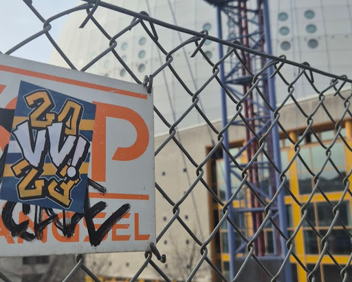 A sticker featuring the text '24 vv!' is placed on a sign, which itself is attached to a chain-link fence. The sticker has a blue background with orange numbers and letters outlined in black. The letters 'vv!' are in a stylized graffiti font. The sign beneath the sticker has a white background with an orange stripe and a large orange letter 'P'. There is also some graffiti tagging on the lower part of the sign. In the background, a building with rounded windows and some steel infrastructure is visible.
