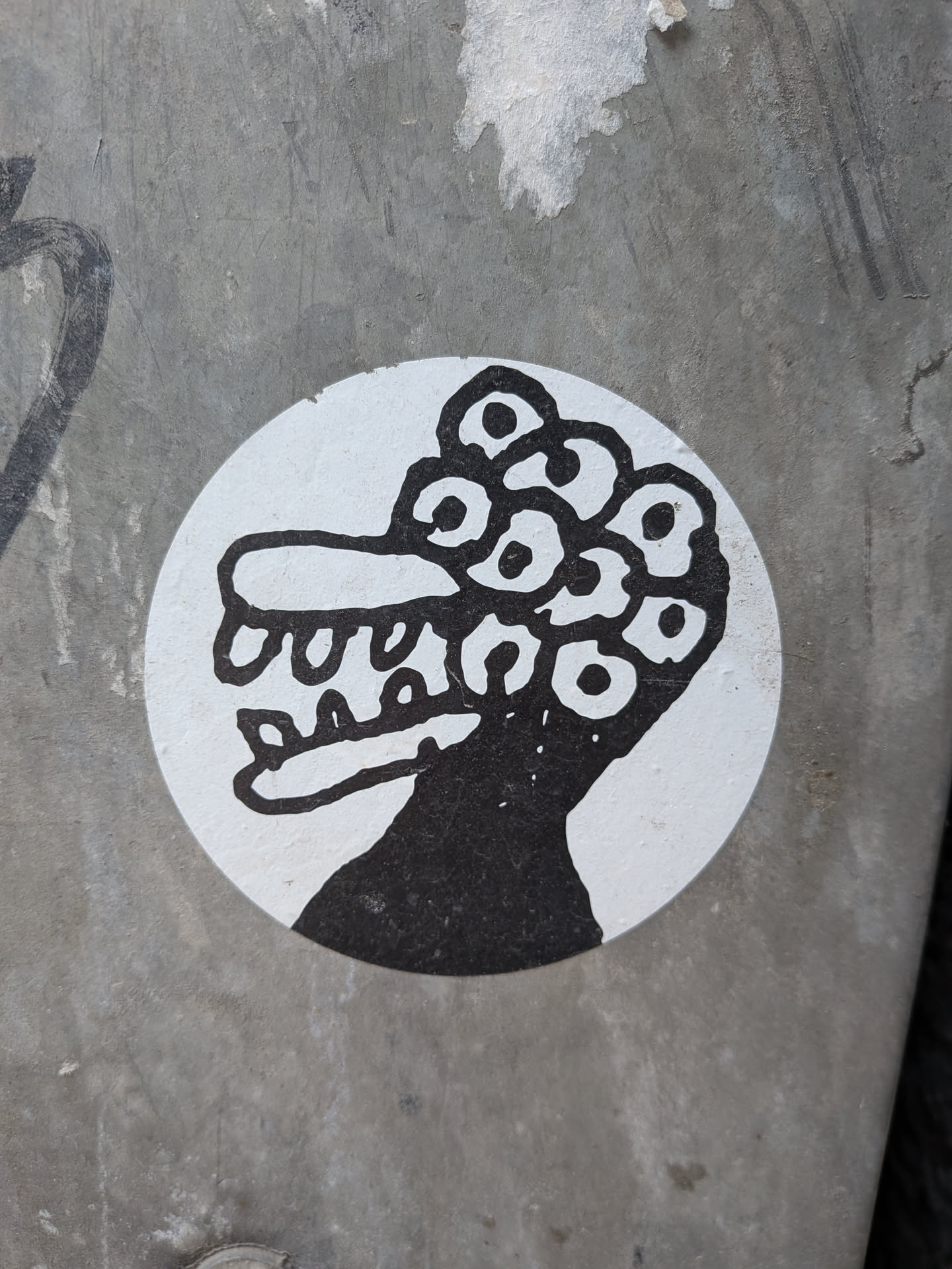 The sticker is circular, featuring a stark black-and-white design. It depicts a stylized, somewhat monstrous or alien-like head in profile, facing left. The creature has a prominent snout or jawline with several rounded, tooth-like protrusions. The most striking feature is the top of its head, which is covered in a dense cluster of numerous circular eyes, each with a black pupil. The creature's body or neck extends downwards, rendered in solid black, contrasting with the white background of the circular sticker. The lines are thick and bold, giving it a cartoonish yet slightly unsettling appearance.
