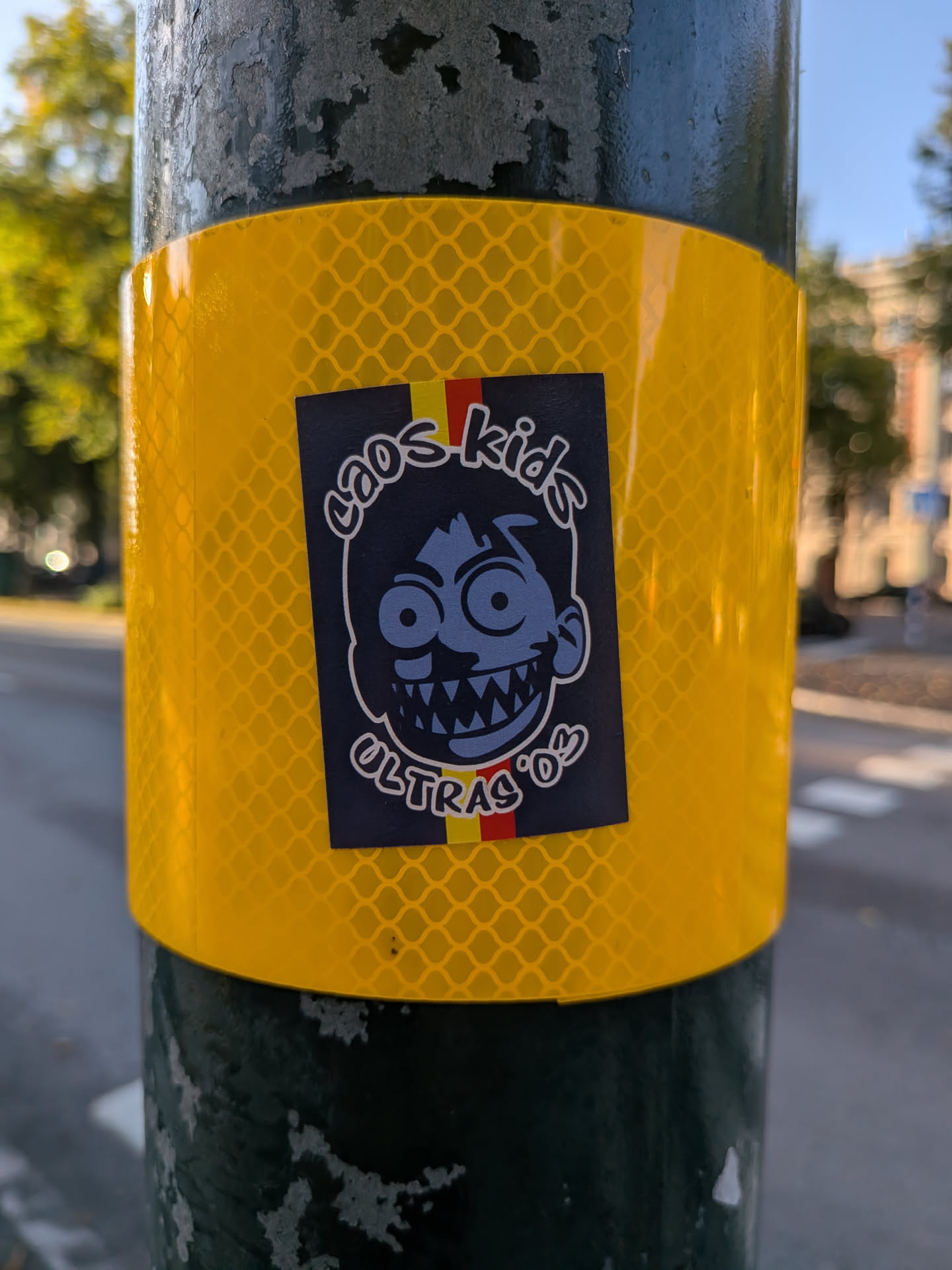 Rectangular sticker on a yellow reflective band. Features a manic, wide-eyed blue cartoon face with sharp teeth. A vertical yellow and red stripe sits behind the character. Text reads 'caos kids' above and 'ULTRAS '03' below in white stylized font.