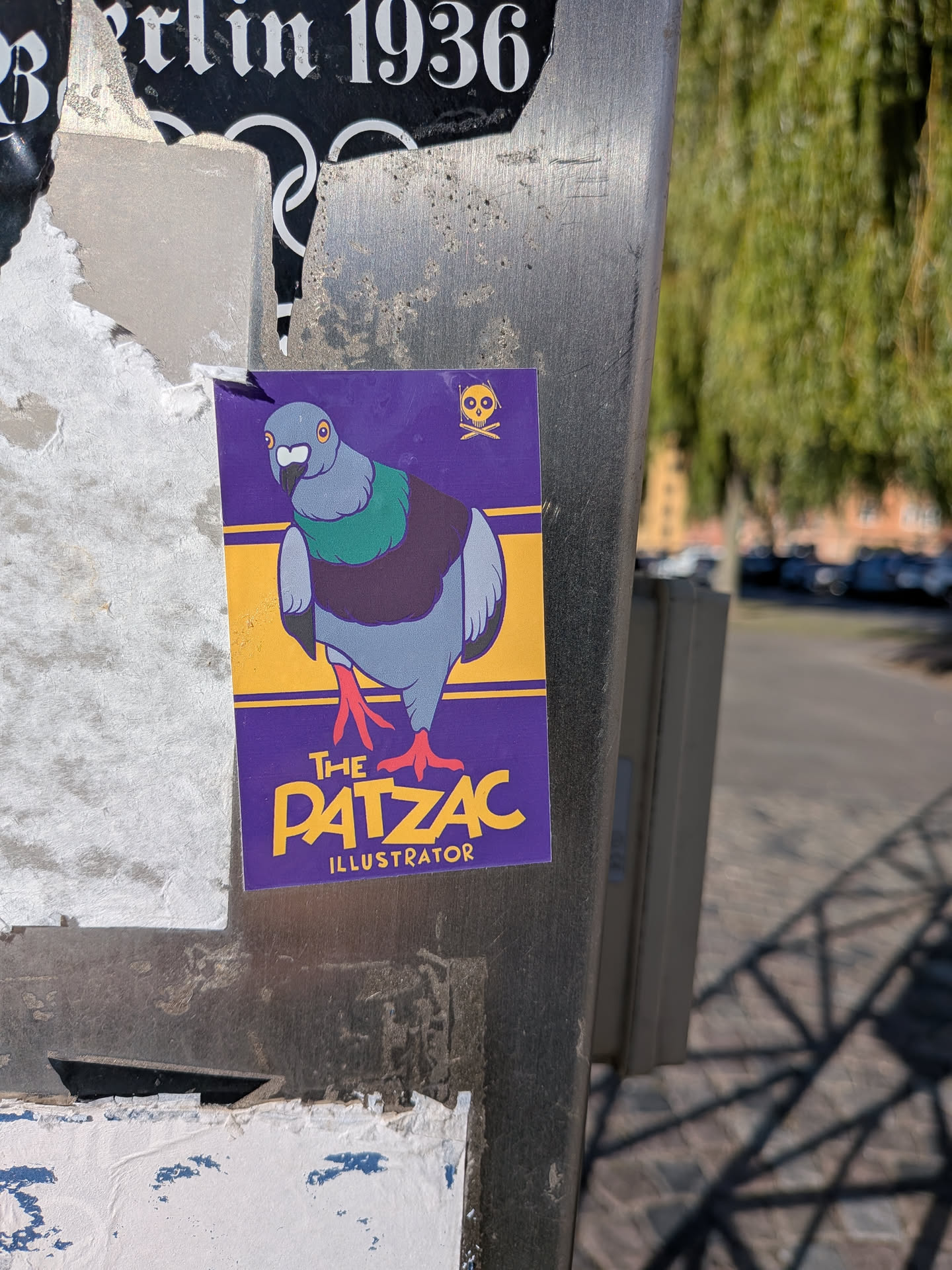 The sticker is predominantly purple with a yellow border. It features a stylized cartoon pigeon with grey and green plumage, large orange eyes, and red feet. Above the pigeon, in the top right corner, is a small yellow skull and crossbones symbol. Below the pigeon, the text 'THE PATZAC ILLUSTRATOR' is displayed in bold yellow lettering, with 'ILLUSTRATOR' in a smaller font.