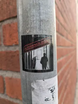 A black and white sticker featuring a person walking down a hallway with the text "Fråga Kriminologen" (Ask the Criminologist) in red and black. The sticker is affixed to a metal pole against a brick wall.