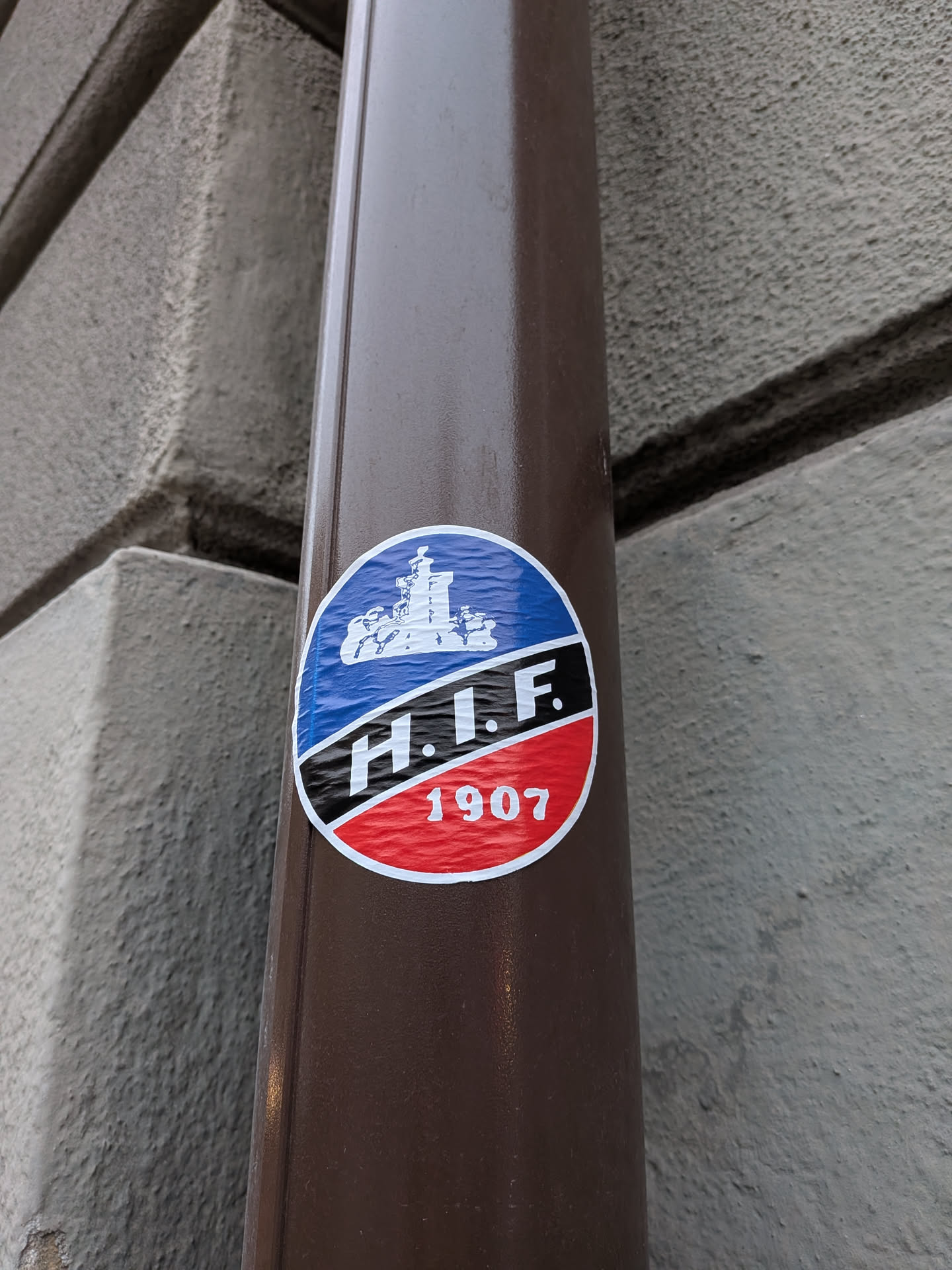 Street sticker The sticker is circular and features a design divided into three horizontal sections. The top section is blue with a white silhouette of a ship with sails, set against a wavy water background. The middle section is black with white, bold sans-serif lettering that reads 'H.I.F.'. The bottom section is red with white numerals '1907'. The entire design is outlined in white.