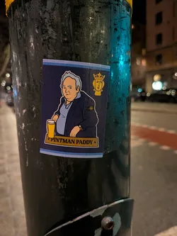 Street sticker The sticker features a cartoon illustration of a portly man with light skin and grey hair, wearing a dark blue jacket over a light blue polo shirt. A distinctive circular badge, resembling the Stone Island compass logo, is visible on his left sleeve. He holds a yellow pint glass filled with a golden beverage. In the top right corner, there's a stylized yellow head wearing a crown. The sticker has a dark blue background with light blue and white horizontal stripes at the bottom. A yellow banner across the bottom reads "PINTMAN PADDY" in dark text, flanked by two yellow stars. The sticker is rectangular with slightly rounded bottom corners.
