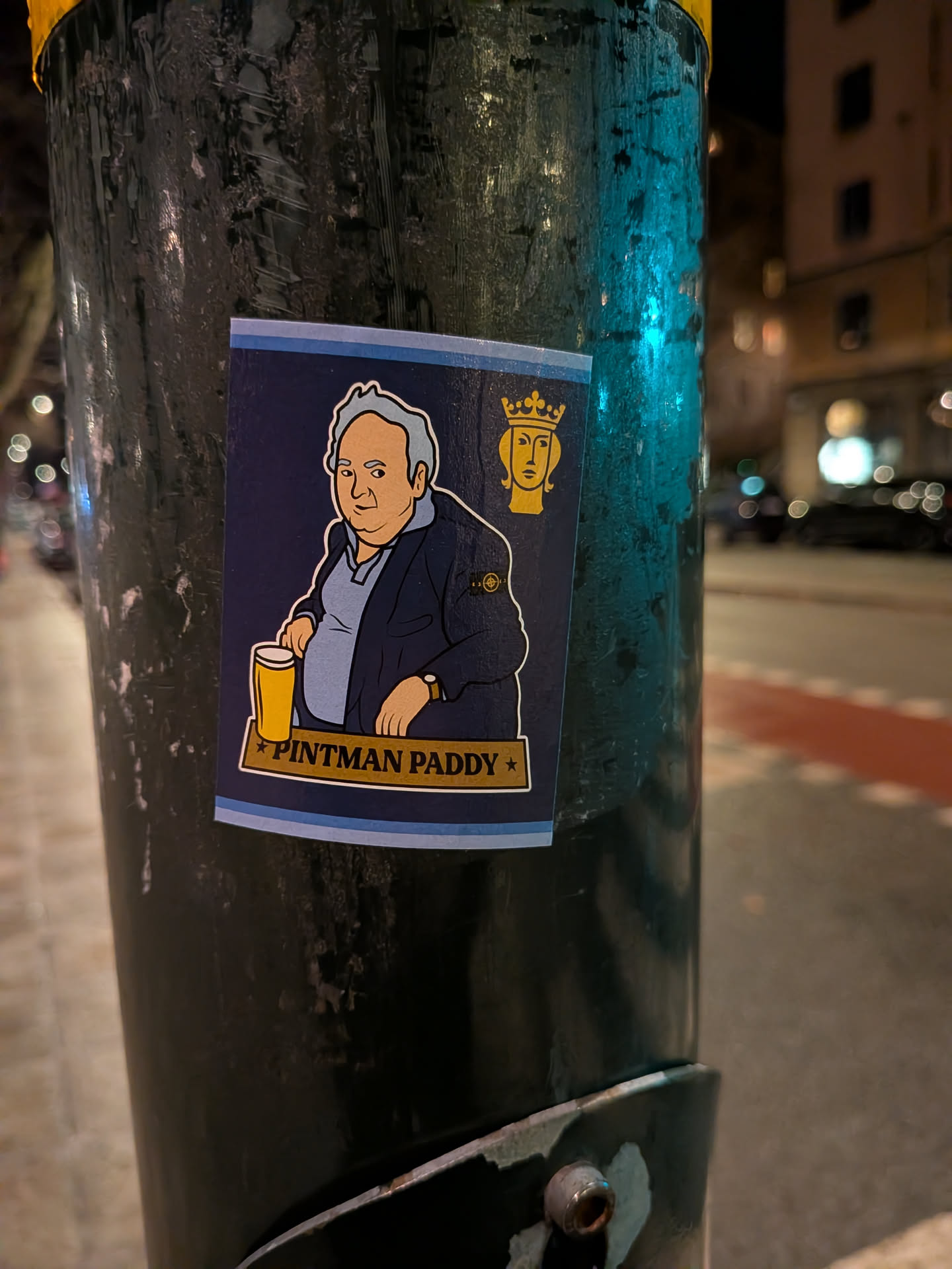 The sticker features a cartoon illustration of a portly man with light skin and grey hair, wearing a dark blue jacket over a light blue polo shirt. A distinctive circular badge, resembling the Stone Island compass logo, is visible on his left sleeve. He holds a yellow pint glass filled with a golden beverage. In the top right corner, there's a stylized yellow head wearing a crown. The sticker has a dark blue background with light blue and white horizontal stripes at the bottom. A yellow banner across the bottom reads "PINTMAN PADDY" in dark text, flanked by two yellow stars. The sticker is rectangular with slightly rounded bottom corners.