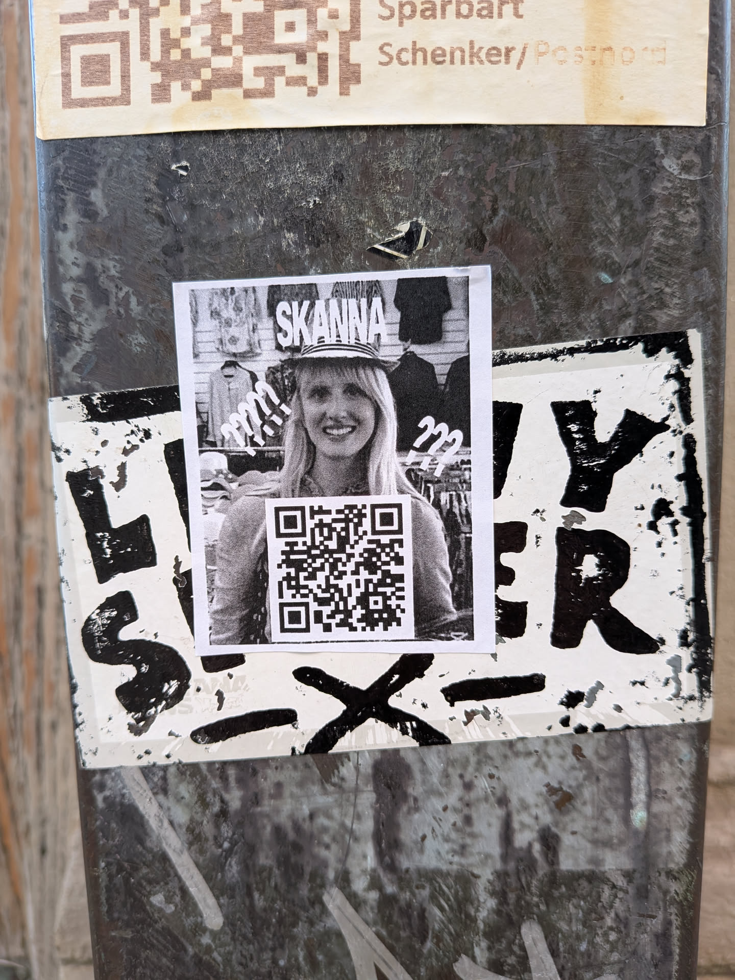 A rectangular monochrome sticker showing a smiling woman in a hat. The Swedish word 'SKANNA' is written above her, surrounded by question marks. A prominent QR code is placed in the center foreground.