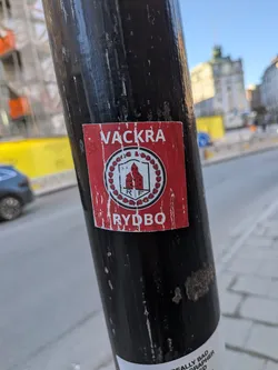 Street sticker A square sticker with a red background, featuring a central circular emblem. The emblem has a white inner circle containing a stylized red building (resembling a church or historic structure) with the letters 'R F' in black below it. This is surrounded by a white ring adorned with a pattern of small red leaf-like or tulip-like shapes, all enclosed by a black border. Above the emblem, in white capital letters, is the word 'VACKRA', and below it, also in white capital letters, is 'RYDBO'. The sticker shows significant wear and tear, with numerous white scuffs and scratches across its surface, indicating its age and exposure to elements.