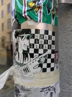 Street sticker The sticker shows a black and white illustration of a musician wearing sunglasses and playing an electric guitar. The background is a checkered pattern. Above the musician, the text "CUMBIA BOYS ÖREBRO" is written in a unique font. The sticker is adhered to a pole or post, with other stickers and remnants of stickers also visible.