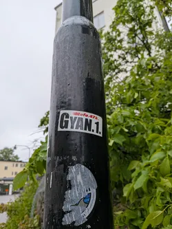 Street sticker A rectangular sticker with a white background and slightly rounded corners, adhered to a dark, weathered pole. The dominant feature is the text 'GYAN.1.' in large, bold, black sans-serif letters. Above and slightly to the left of 'GYAN.1.', smaller text reads 'Moto.47k.' in a stylized red font, which appears to have a subtle flame or speed-like graphic element. The sticker shows minor signs of wear, particularly around its edges.