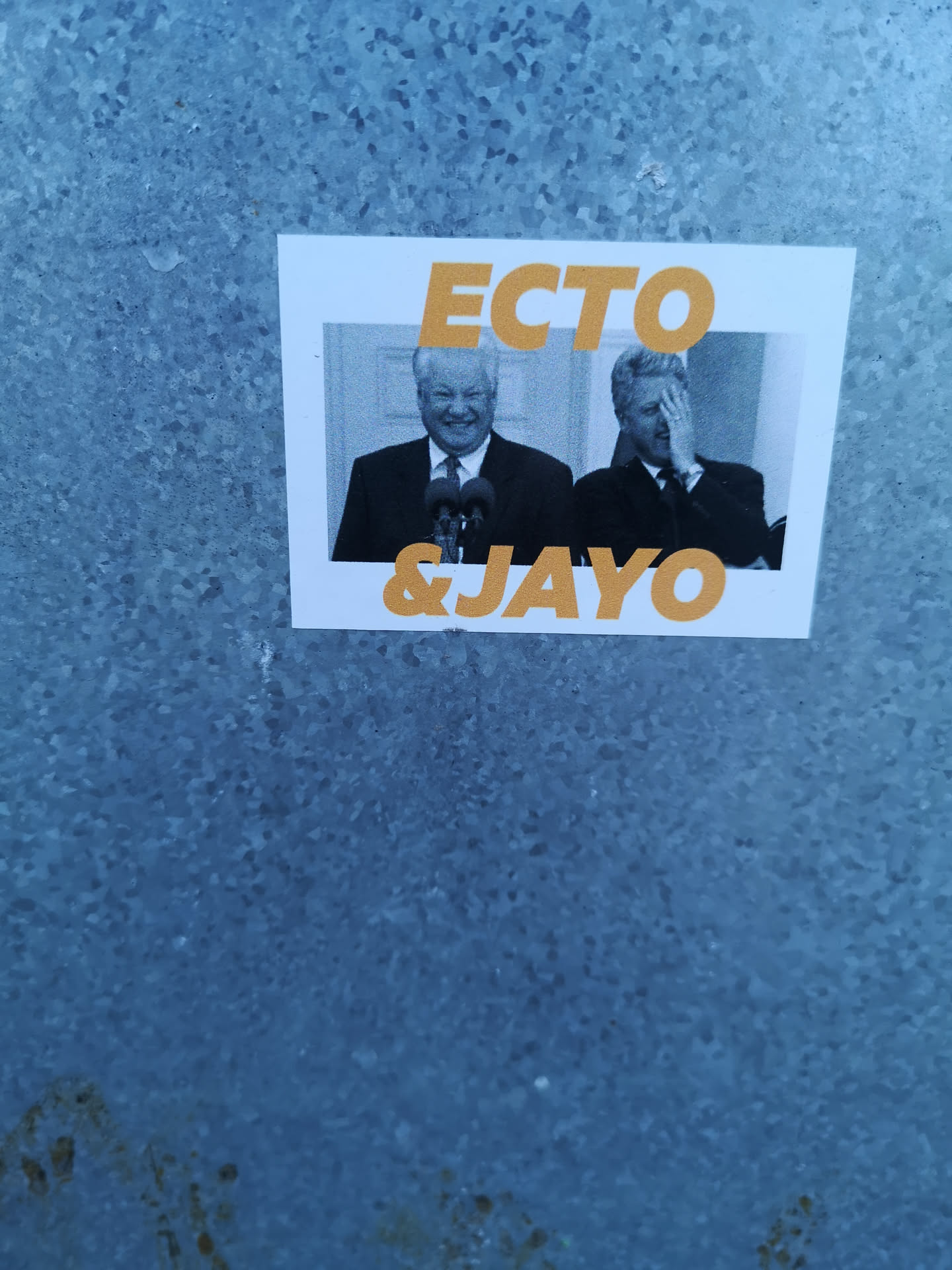 A rectangular sticker featuring a black and white photograph of two prominent political figures. On the left, Boris Yeltsin is seen smiling broadly behind microphones. On the right, Bill Clinton is shown covering his face with his hand. The photograph has a white border. Overlaying the image, in a bold, sans-serif, orange-yellow font, are the words 'ECTO' at the top and '& JAYO' at the bottom. The sticker is adhered to a textured, blue-grey metallic surface.