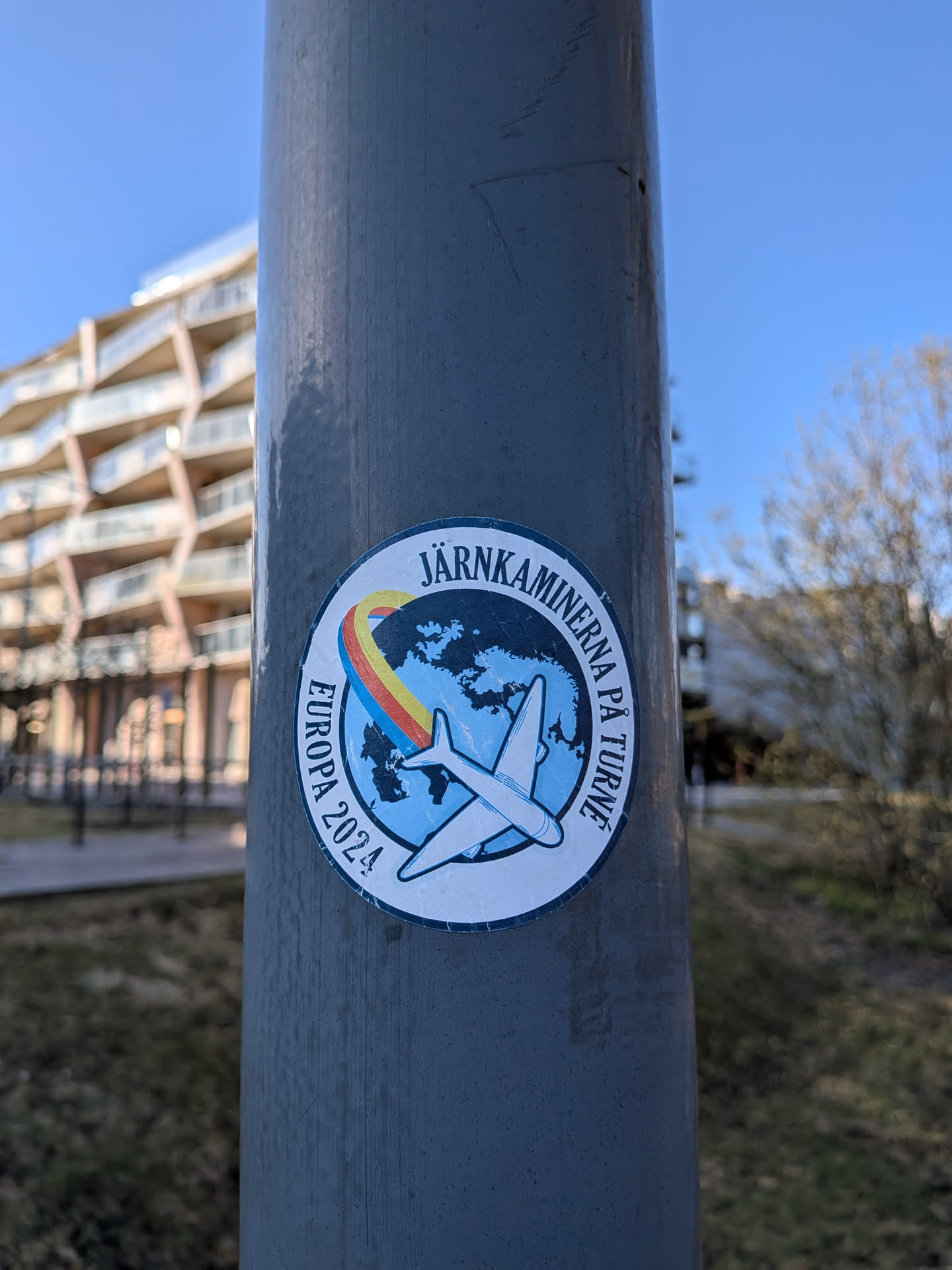 A circular sticker featuring a light blue globe with darker blue landmasses at its center. A white airplane with a black outline is depicted flying across the globe, leaving a vibrant, arcing contrail in shades of red, yellow, and green. The top arc of the sticker contains the text "JÄRNKAMINERNA PÅ TURNÉ" in black capital letters, while the left side features "EUROPA 2024" also in black capital letters, all set against a white outer ring.