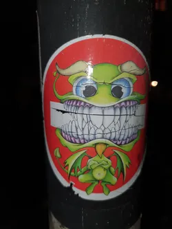 Street sticker This sticker features a cartoonish green devil with large eyes, horns, and wings. It's got a big toothy grin, and its arms are crossed. The sticker has a red circle background and is slightly damaged around the edges.
