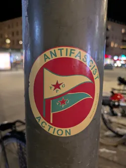 Street sticker The sticker is round, with a cream-colored background and a red circle in the center. Inside the circle are two flags: the top flag is gold with a red star and the letters 'YPG', and the bottom flag is teal with a red star and the letters 'YPJ'. The text 'ANTIFASCIST ACTION' curves around the top and bottom of the circle in green. Next to the flags it reads 'VRIJEBOND.ORG'.