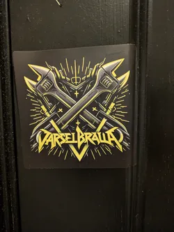 The sticker is predominantly black with vibrant yellow and metallic gray accents. At its center are two crossed adjustable wrenches, depicted with a metallic sheen. Behind and around the wrenches, stylized yellow lines radiate outwards, resembling bursts of energy or light. The overall design has a strong, industrial, and somewhat aggressive feel. Below the wrenches, bold, stylized yellow lettering spells out "VARSELBRAVA", with sharp angles and a metallic texture. Small cross symbols are subtly integrated into the design, adding to its symbolic nature.