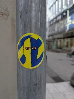 Street sticker The sticker is circular, featuring a stylized head wearing a balaclava or ski mask. The face is vertically split down the middle, with the left half (from the viewer's perspective) being yellow and the right half blue. The eyes are the only visible facial features, rendered with some detail, showing brown irises and dark pupils. The background behind the head is also split diagonally, with a yellow lightning bolt or diagonal stripe on the blue side, and a blue lightning bolt or diagonal stripe on the yellow side, creating a dynamic 'X' or cross pattern. The entire design is outlined in black, and the sticker itself shows minor wear and tear, particularly along its edges, indicating it's a physical sticker adhered to a metal pole.