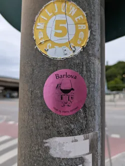 Street sticker The central sticker is circular and bright pink (magenta). At the top, the word "Barlova" is written in black text. Below it, there is an intricate black line drawing of a wine glass, within the base and stem of which is depicted a detailed illustration of a building or house. Along the bottom curve of the sticker, the address "TÄHE 29, TARTU, ESTONIA" is printed, indicating a specific location in Tartu, Estonia. The sticker is affixed to a weathered grey pole.