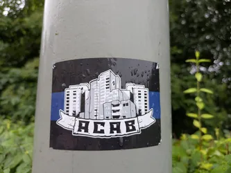 Street sticker The sticker is rectangular with slightly rounded corners, adhered to a light grey pole. Its background is dark grey or black, covered with numerous small water droplets. Across the center, a horizontal blue stripe is visible. Overlaid on this stripe and the dark background is an illustration of a cluster of buildings in varying shades of grey and white, giving a monochrome, almost sketched appearance. These buildings are stylized, resembling apartment blocks or high-rises. Below the cityscape, a white banner with jagged ends unfurls, containing the bold, blocky text "ACAB" in black. The entire illustration (buildings and banner) has a white outline, making it stand out from the dark background. The sticker appears slightly worn at the top right corner.