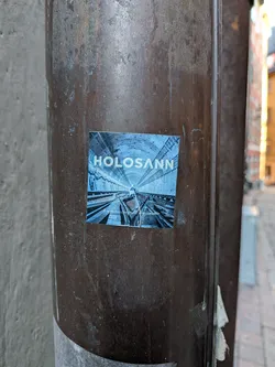 A square sticker featuring a striking image of a long, illuminated tunnel with train tracks receding into the distance. The perspective is central, drawing the eye deep into the tunnel. The dominant colors are cool blues and grays, giving the scene a somewhat futuristic or mysterious ambiance. The tunnel walls show concrete segments and some visible wiring or pipes. In the upper center, the word "HOLOSANN" is prominently displayed in white, sans-serif capital letters. Below the image, near the bottom edge, smaller white text reads "A la recherche d'une ville parallèle."