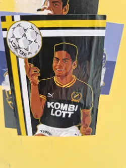 Street sticker A stylish sticker showcasing a young Black man with a 1980s or 1990s haircut, wearing a black and gold football jersey with the text "KOMBI LOTT" printed on it. He's depicted juggling a football. The sticker has a black background with yellow and white accents.