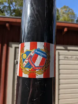 Street sticker Rectangular sticker with red and white vertical stripes. Features overlapping crests of Lyn Fotball and Bastionen enclosed in a golden laurel wreath. A red ribbon at the bottom contains Norwegian text.
