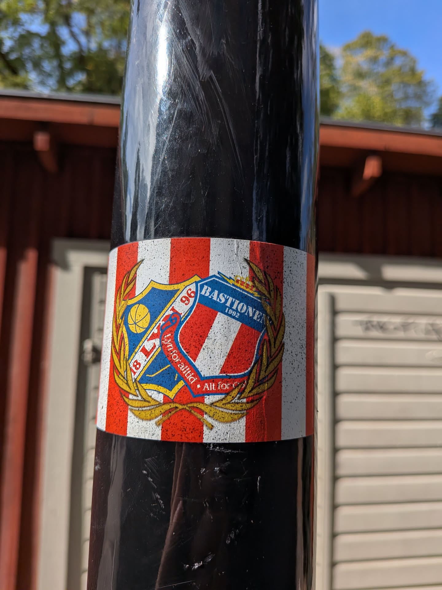 Rectangular sticker with red and white vertical stripes. Features overlapping crests of Lyn Fotball and Bastionen enclosed in a golden laurel wreath. A red ribbon at the bottom contains Norwegian text.