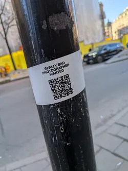 Street sticker A white rectangular sticker wrapped around a weathered, black metal pole. The sticker features bold, black, uppercase text that reads 'REALLY BAD PHOTOGRAPHER WANTED' in a sans-serif font. Below the text, a prominent black and white QR code is centered. The sticker appears slightly creased where it conforms to the pole's curvature.