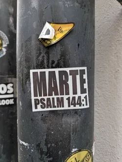Street sticker A rectangular sticker with a white background and dark, bold, sans-serif text. The top line prominently displays 'MARTE' in large uppercase letters, while the bottom line reads 'PSALM 144:1' in slightly smaller uppercase letters. The sticker is affixed to a dark, textured pole, showing some minor wear and tear.