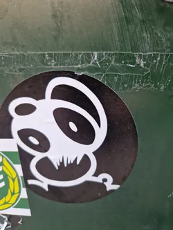 Street sticker A round black and white sticker featuring a stylized panda design. The panda is depicted in a playful, cartoonish style with simple lines and bold features. The sticker is affixed to a dark green surface that shows signs of wear and tear.