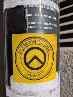 The sticker is square-shaped with slightly rounded corners, featuring a vibrant yellow background. At its center is a prominent circular design in black. This black circle contains a thick outer ring and a central symbol: a black circle with a thick, upward-pointing chevron or arrow inside, resembling a 'no entry' sign with an added element. Arcing along the top inner edge of the black circle is the text 'GENERATION IDENTITÆR' in black capital letters, and similarly, 'DANMARK' is arced along the bottom. Below the main circular emblem, on the yellow background, the website 'identitaer.dk' is printed in smaller black lowercase letters. The sticker shows signs of wear, with slight wrinkles and minor tears, indicating it has been affixed for some time on a dark, possibly black, pole.