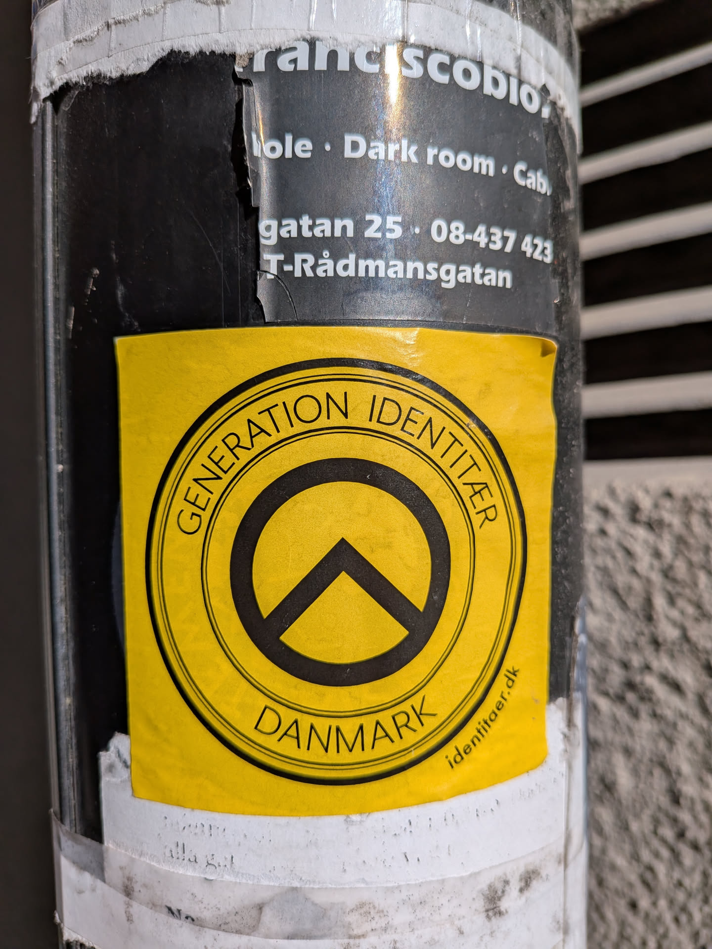 The sticker is square-shaped with slightly rounded corners, featuring a vibrant yellow background. At its center is a prominent circular design in black. This black circle contains a thick outer ring and a central symbol: a black circle with a thick, upward-pointing chevron or arrow inside, resembling a 'no entry' sign with an added element. Arcing along the top inner edge of the black circle is the text 'GENERATION IDENTITÆR' in black capital letters, and similarly, 'DANMARK' is arced along the bottom. Below the main circular emblem, on the yellow background, the website 'identitaer.dk' is printed in smaller black lowercase letters. The sticker shows signs of wear, with slight wrinkles and minor tears, indicating it has been affixed for some time on a dark, possibly black, pole.