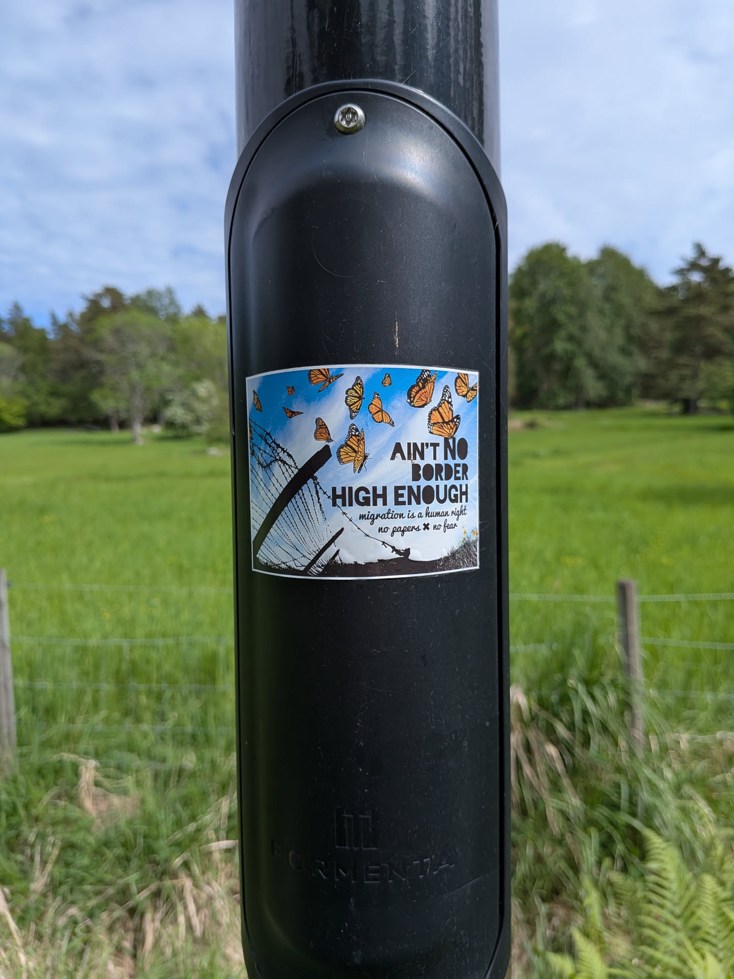 A rectangular sticker showing orange monarch butterflies flying over a barbed-wire fence silhouette against a blue sky. Text on the right reads 'AIN'T NO BORDER HIGH ENOUGH' with smaller subtext supporting migration rights.