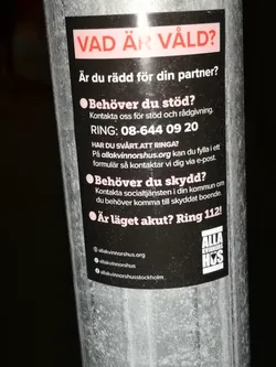 A rectangular, vertically oriented sticker with a black background, adhered to a metal pole. At the top, a light pink/white banner features the question 'VAD ÄR VÅLD?' (WHAT IS VIOLENCE?) in bold red text. Below this, the sticker presents information and questions in white text, such as 'Är du rädd för din partner?' (Are you afraid of your partner?), 'Behöver du stöd?' (Do you need support?), and 'Behöver du skydd?' (Do you need protection?). It provides a phone number '08-644 09 20', a website 'allakvinnorshus.org', and an emergency number 'Ring 112!'. The bottom left corner lists social media handles and a website, indicated by small icons (globe, Instagram, Facebook). In the bottom right, there is a white logo for 'ALLA KVINNORS HUS' (All Women's House) with stylized text.
