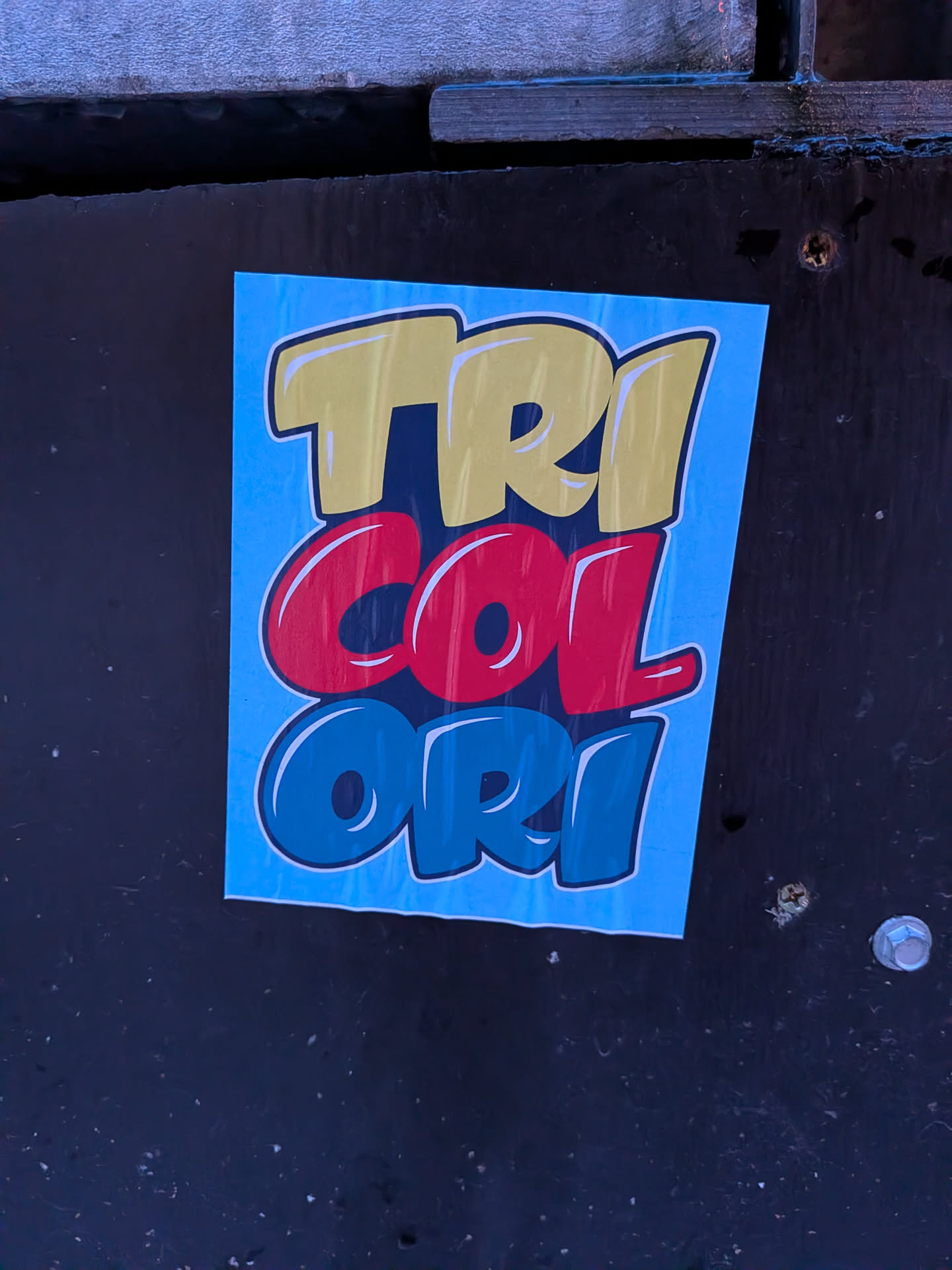 A rectangular sticker featuring the word 'TRICOLORI' stacked vertically on a light blue background. The letters have a bold, cartoonish, or bubble-letter style with white highlights and a dark blue/black outline, giving them a glossy, three-dimensional appearance. The word is split into three parts, each in a distinct color: 'TRI' is yellow, 'COL' is red, and 'ORI' is blue. The sticker is applied to a dark, textured surface, showing slight creasing on its left side.