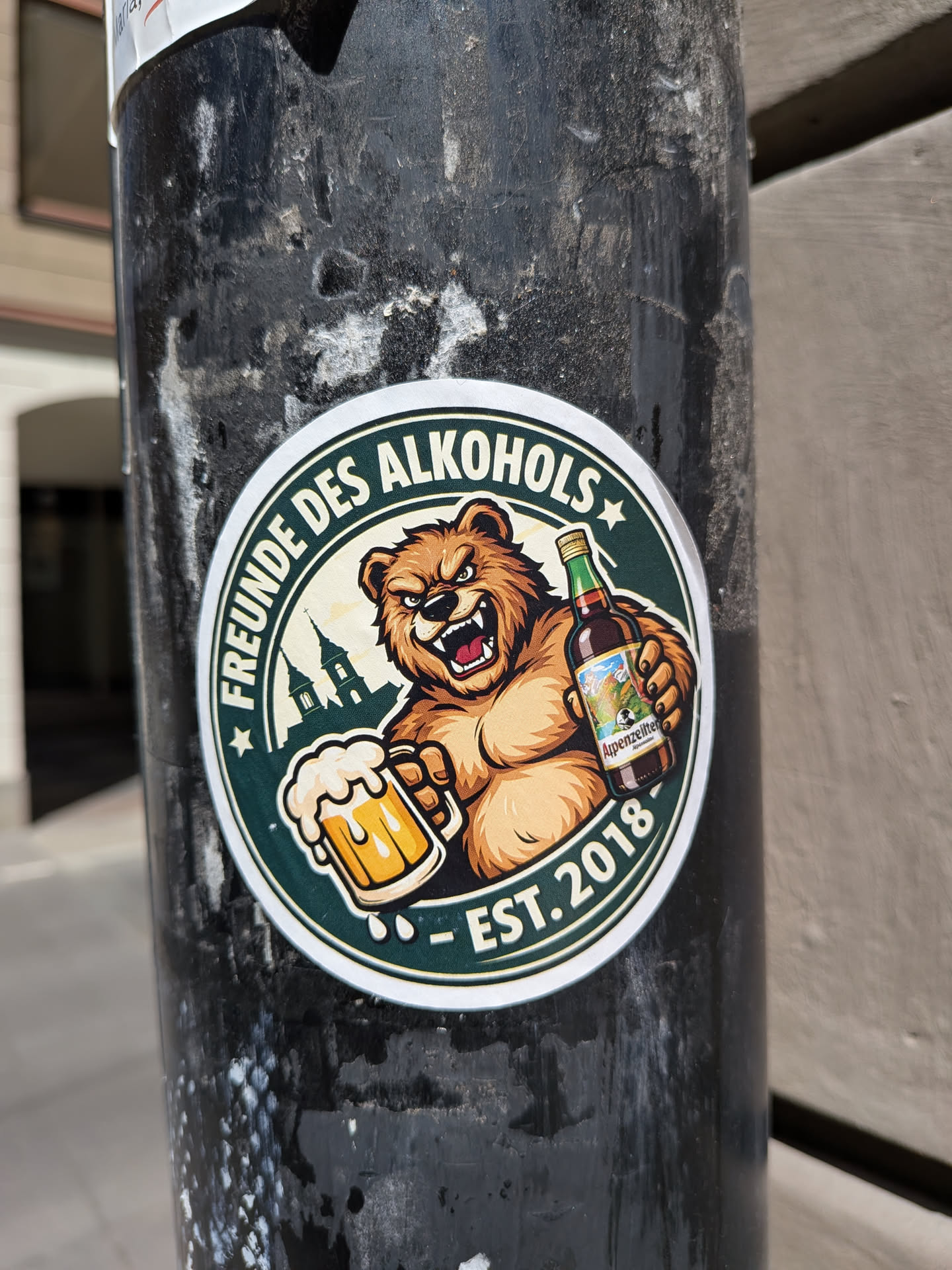 Circular sticker with a dark green border. Features an aggressive brown bear holding a foaming beer mug and an Appenzeller beer bottle. Includes a silhouette of church towers and German text.