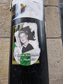 Street sticker Circular sticker with green and white stripes. Features a black-and-white photo of a person with glasses and a headband. Swedish text 'VI KÄKAR DJURGÅRN TILL FRUKOST' is printed at the bottom.