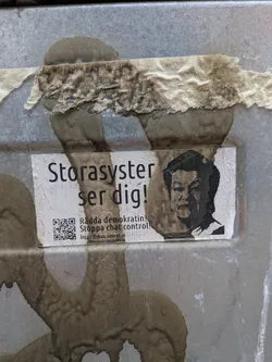A rectangular sticker, predominantly white with black text and imagery, affixed to a weathered, grey metallic surface. The sticker itself shows signs of wear and water damage, with some discoloration and faint stains. On the right side, there is a black and white, somewhat pixelated portrait of an Asian man, resembling a political figure, with a serious expression. To the left of the portrait, bold black text in Swedish reads 'Storasyster ser dig!' (Big Sister is watching you!). Below this, smaller text states 'Rädda demokratin! Stoppa chat control!' (Save democracy! Stop chat control!). In the bottom left corner, a clear black and white QR code is visible, accompanied by the URL 'https://chatcontrol.se'. The overall aesthetic is stark and activist-oriented.