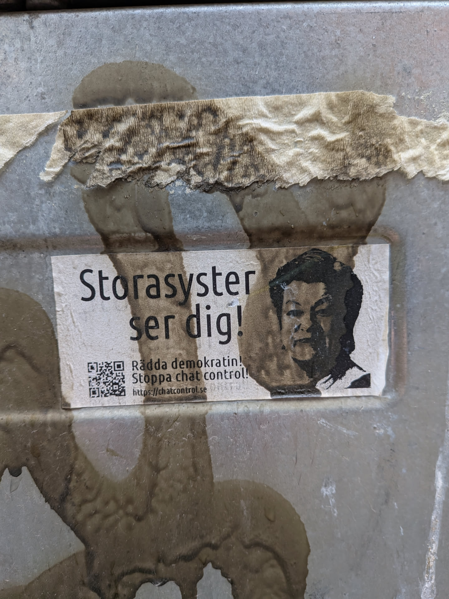 A rectangular sticker, predominantly white with black text and imagery, affixed to a weathered, grey metallic surface. The sticker itself shows signs of wear and water damage, with some discoloration and faint stains. On the right side, there is a black and white, somewhat pixelated portrait of an Asian man, resembling a political figure, with a serious expression. To the left of the portrait, bold black text in Swedish reads 'Storasyster ser dig!' (Big Sister is watching you!). Below this, smaller text states 'Rädda demokratin! Stoppa chat control!' (Save democracy! Stop chat control!). In the bottom left corner, a clear black and white QR code is visible, accompanied by the URL 'https://chatcontrol.se'. The overall aesthetic is stark and activist-oriented.